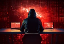 a hooded hacker sitting in front of a desk with two laptops on it